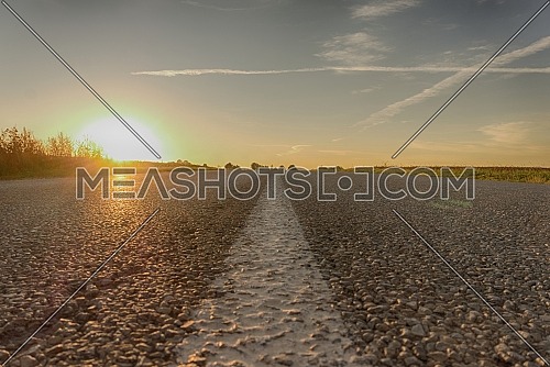 Ground level view of an asphalt road in the countryside at sunset with a view down the receding white center line to a colorful orange sky with lens flare