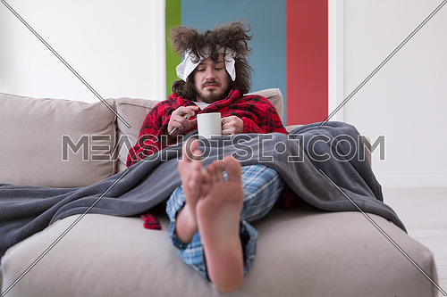 young Man in bathrobe with flu and fever wrapped holding cup of healing tea while sitting on sofa at home