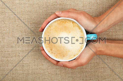 Two woman hands holding embracing big latte cappuccino coffee blue cup over linen canvas background