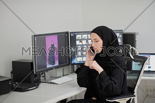 Muslim female graphic designer talking on the phone. Freelancer editing photos on two monitors. Hijab girl on the work place