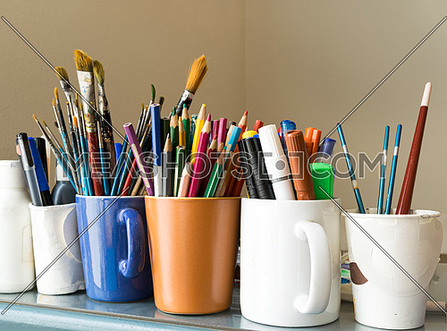 Close up of different used paint brushes, sharpened colored pencils, pens, and markers on colored mugs over blue table and beige background