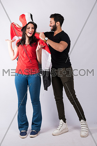 young couple standing and cheering holding big egyptian flag, while the female wearing egyptian flag hat on white background