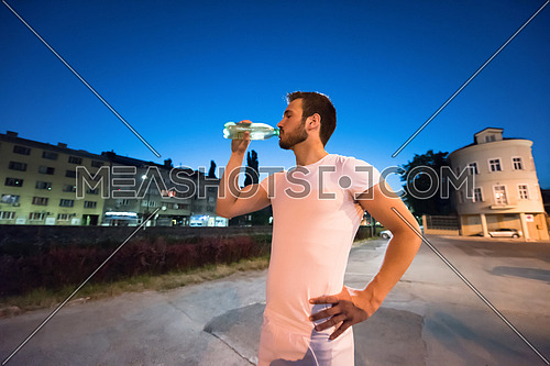 young athletic man drinking water after a night running session in the city