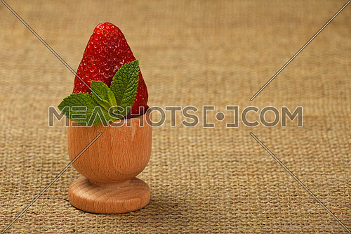 One big red mellow strawberry and green fresh mint leaves in wooden eggcup holder on natural burlap jute canvas background