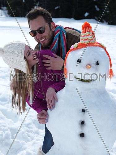 portrait of happy young couple with snowman at beautiful winter day with fresh snow and forest in background