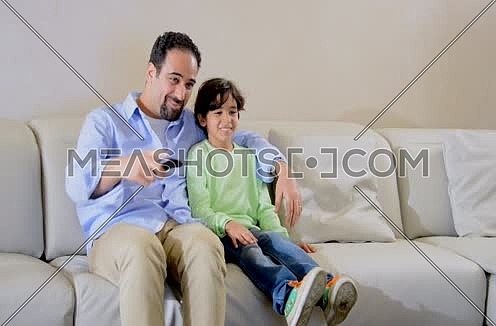 Mid shot for a male watching TV holding remot control and his son come to watch with him