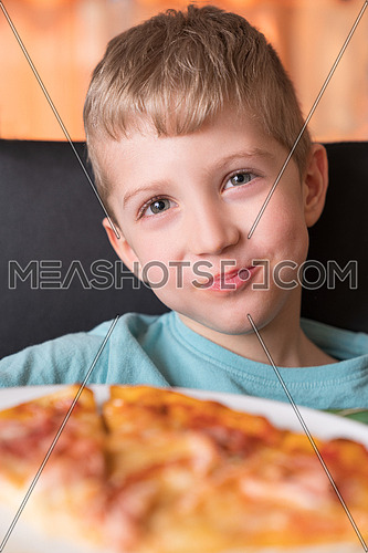 Beautiful happy young boy smiling and eating fresh made pizza,She sit at black chair, He has blonde hair.