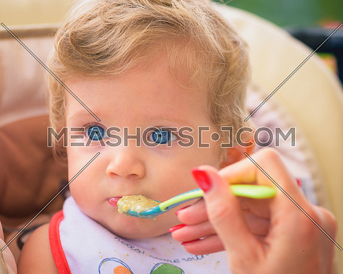 A mother feeding her baby boy with spoon outdoor.