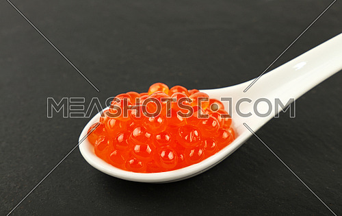 Close up one portion of salmon fish red caviar in white spoon on background of black slate kitchen board, high angle view