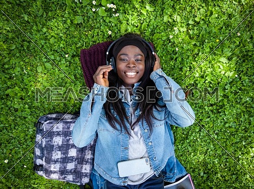 Top view of smiling african woman lying on green grass listening to music with headphones.