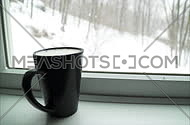 View of a black coffee cup in a window ledge as a winter snow storm blows outside.