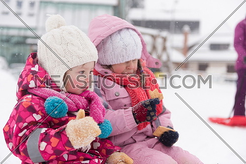 portrait of two cute little grils sitting together on sledges outdoors at snowy winter day, eating tasty cookies on break