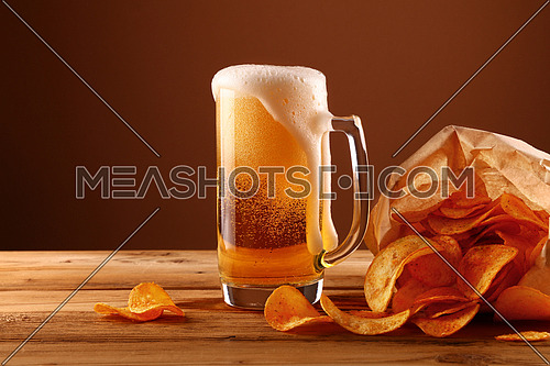 Close up one glass mug of lager beer with white froth and bubbles and paper bag of potato chips on wooden table over dark brown background with copy space, low angle side view