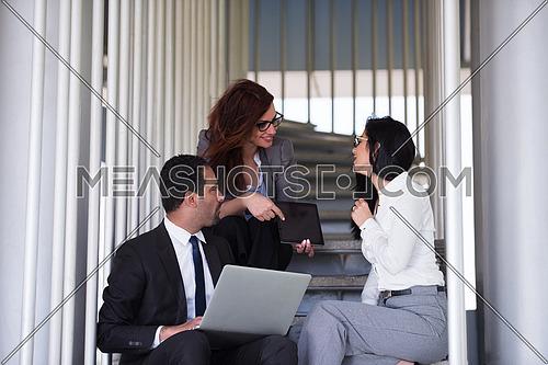 Young executives sitting on the steps of a corporate office building discussing business using laptop and tablet
