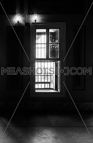 Black and white high contrast shot of narrow window, revealing strong light into dark room with illuminated lanterns and white tiled marble floor