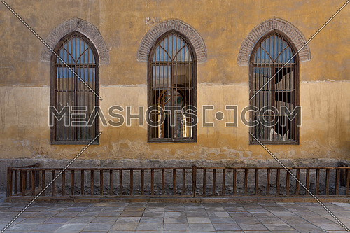 Row of three adjacent wooden grunge arched window with decorated wrought iron grid over yellow stone bricks wall and wooden balustrade, Medieval Cairo, Egypt