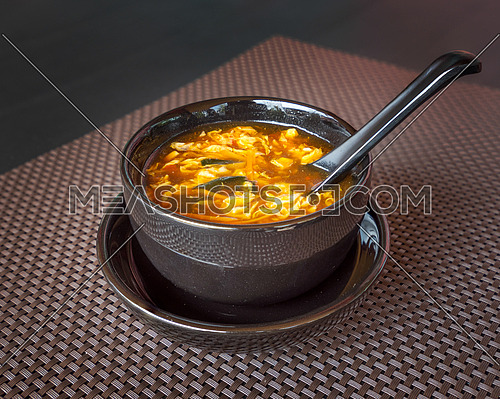 Chinese food, hot and sour soup served in black bowl with spoon on dark background.