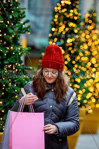 Happy young girl with shopping bags walking along city street, colorful lights bokeh background