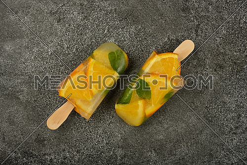 Close up two fruit ice cream popsicles with fresh orange slices and green mint leaves on gray table surface, elevated top view, directly above