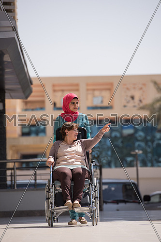 older sick woman in wheelchair with young middle eastern  nurse in front of a large modern hospital