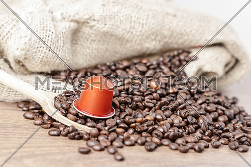 In the foreground a coffee capsule on wooden spoon and  roasted coffee beans with burlap sack on blur wooden background ,close up.