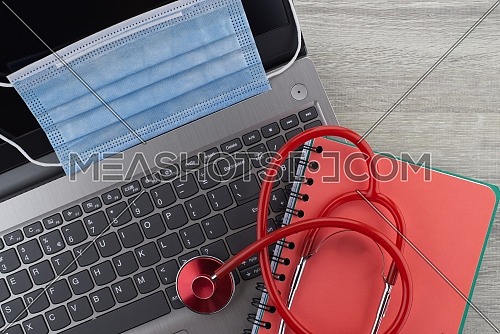 Coronavirus or Covid-19 medical concept with a colorful red stethoscope coiled over a surgical face mask alongside a notebook and pen on a wooden desk with computer keyboard viewed top down