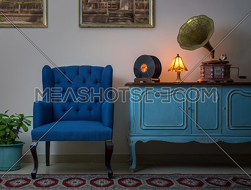 Interior shot of blue armchair, vintage wooden light blue sideboard, lighted antique table lamp, old phonograph (gramophone), vinyl records on background of beige wall and tiled porcelain floor