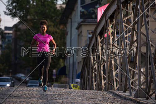 Young sporty african american woman running on sidewalk across the bridge at early morning jogging with city sunrise scene in background