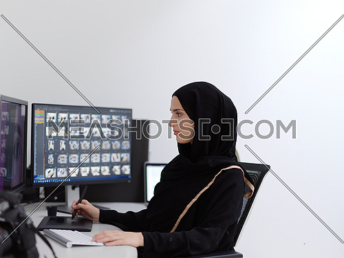 Muslim female graphic designer working on computer using graphic tablet and two monitors. Girl wearing hijab editing photos in the office