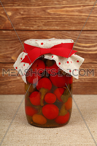 Close up of one glass jar of pickled small round red hot cherry chili peppers with linen canvas lid decoration and red ribbon over brown wooden background, low angle side view