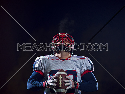 portrait of confident American football player holding ball while standing on field at night