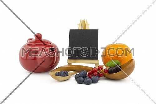 Fresh seasonal fruit still life with small chalkboard and assorted berries including blueberries, cherries and red currants on wooden spoons with orange and colorful red teapot