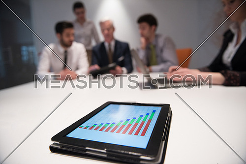 Close up of tablet touchpad computer with focus on business analytics and progress chart document. Business people group on meeting in background