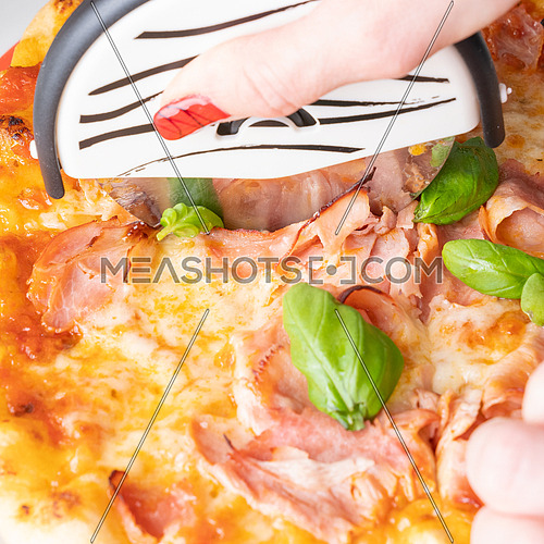 Slicing fresh pizza with cheese(mozzarella), tomato, ham and small fresh basil leaves with special roller knife. Woman hand holding roller knife.