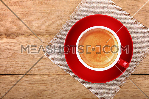 Full big cup of espresso coffee with brown crema froth on red porcelain saucer over wooden table with textile tablecloth napkin, close up, elevated top view, directly above
