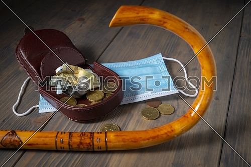 Money spilling from an open purse onto a wooden table with cane and surgical mask alongside viewed from above, old age and pandemic conceptual flat lay still life