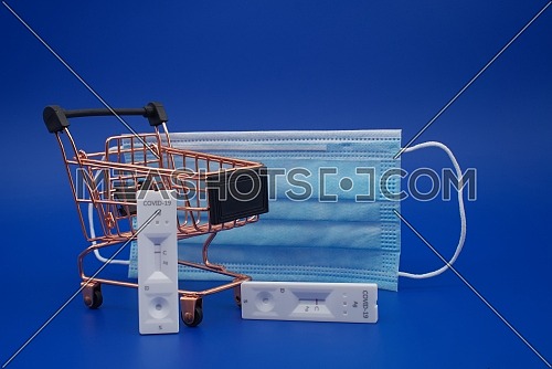 Covid-19 and coronavirus healthcare and shopping concept with mask, shopping cart and rapid antibody test kits over a blue background