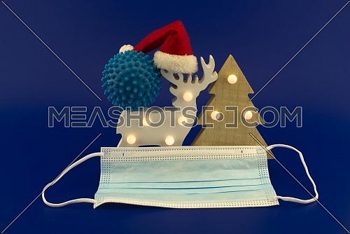 Christmas in pandemic concept still life with blue virus molecule, red Santa hat, Christmas tree, gift box and surgical face mask all isolated on blue background
