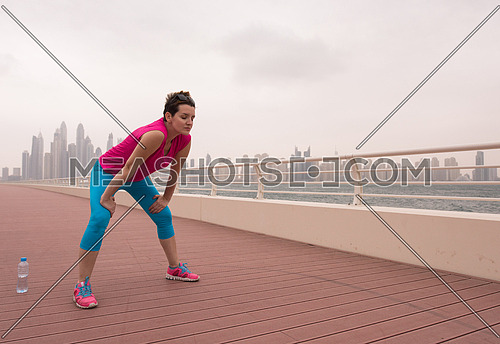 very active young beautiful woman stretching and warming up on the promenade along the ocean side with a big modern city in the background to keep up her fitness levels as much as possible