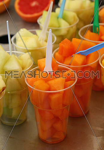 Street food, selection of fruit salads, slices of cut fresh ripe yellow and orange cantaloupe melon cubes in plastic cups with forks at retail market stall display, close up, high angle view