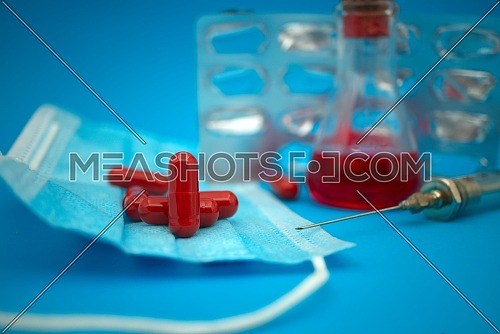 Medical, healthcare or Covid-19 still life with red capsules, a pharmacy bottle, hypodermic syringe, mask and chemical solution in a flasks over a blue background