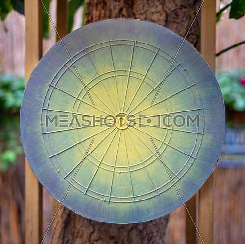 Dartboard game board painted in green, yellow and blue hanged on tree with blurred background of public garden