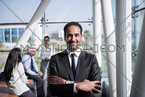 Portrait of a male executive wearing eye glasses and a group meeting is taking place at the background in a bright office