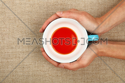 Two woman hands holding embracing big empty finished blue cup of black red tea over linen canvas background, warming hands
