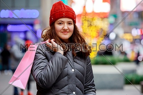 Happy girl with long brown hair standing with shopping bags and portrait, smiling, walking down the street ,colorful lights bokeh background