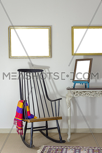 Interior shot of classic rocking chair and wooden ornate brown desktop photo frame on old style vintage table on background of off white wall with two hanged paintings with clipping path for paintings