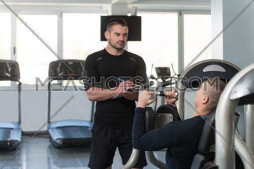 Personal Trainer Takes Notes While Young Man Exercise Chest On Machine In The Gym