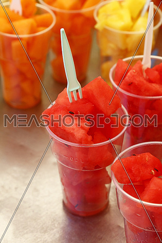 Street food, selection of fruit salads, slices of cut fresh ripe watermelon and melon cubes in plastic cups with forks at retail market stall display, close up, high angle view