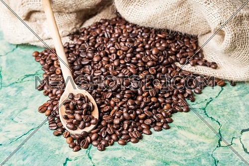 Coffee beans in coffee burlap bag on green table and wooden spoon with coffee beans on top.