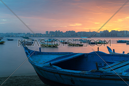 Turquoise blue fishing boat on the beach at sunrise with Alexandria skyline in far distance and colorful sky at sunrise